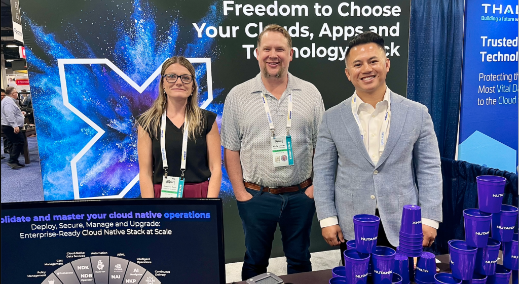 Three professionals at a technology expo booth, smiling in front of a blue space-themed backdrop with lanyards and purple branded cups on the table behind them.