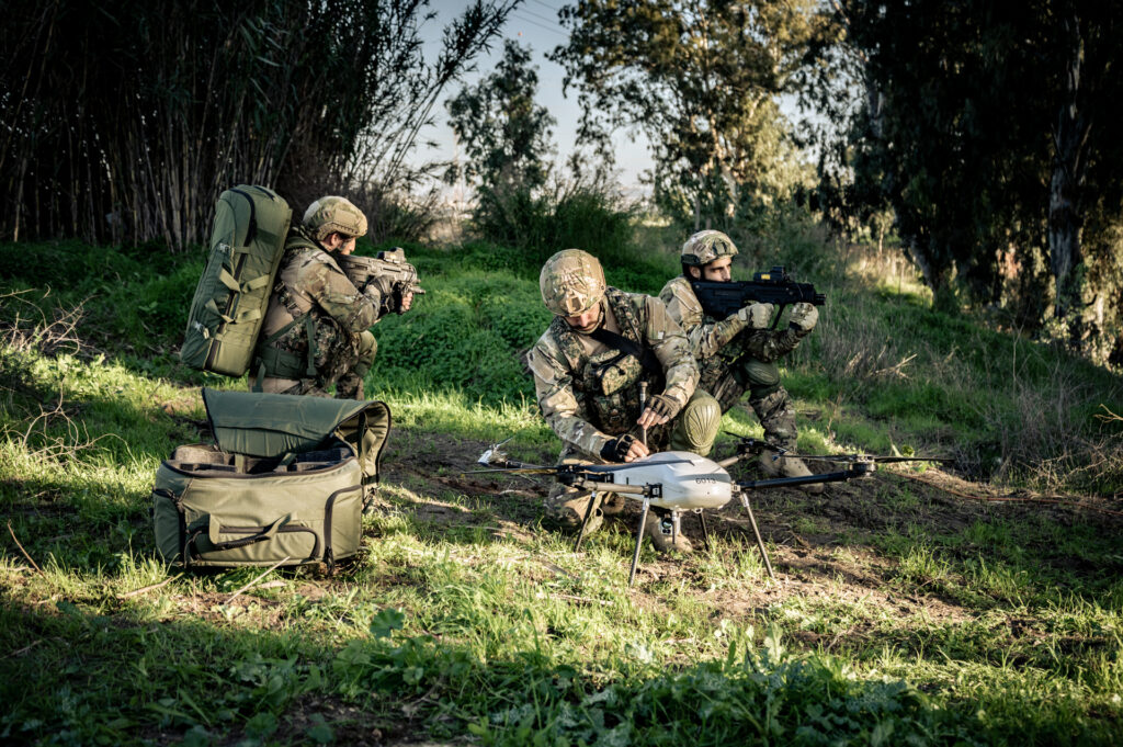 Soldiers in camouflage gear crouch in a field with a white quadcopter-style drone on the ground, packing equipment nearby.