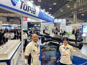 Exhibition hall with attendees at a tech trade show; two staff in white uniforms near a blue Topia booth and a helicopter display in the foreground.