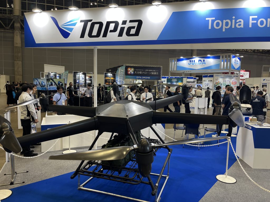 Exhibition hall with a large Topia banner overhead and people viewing drone displays below.