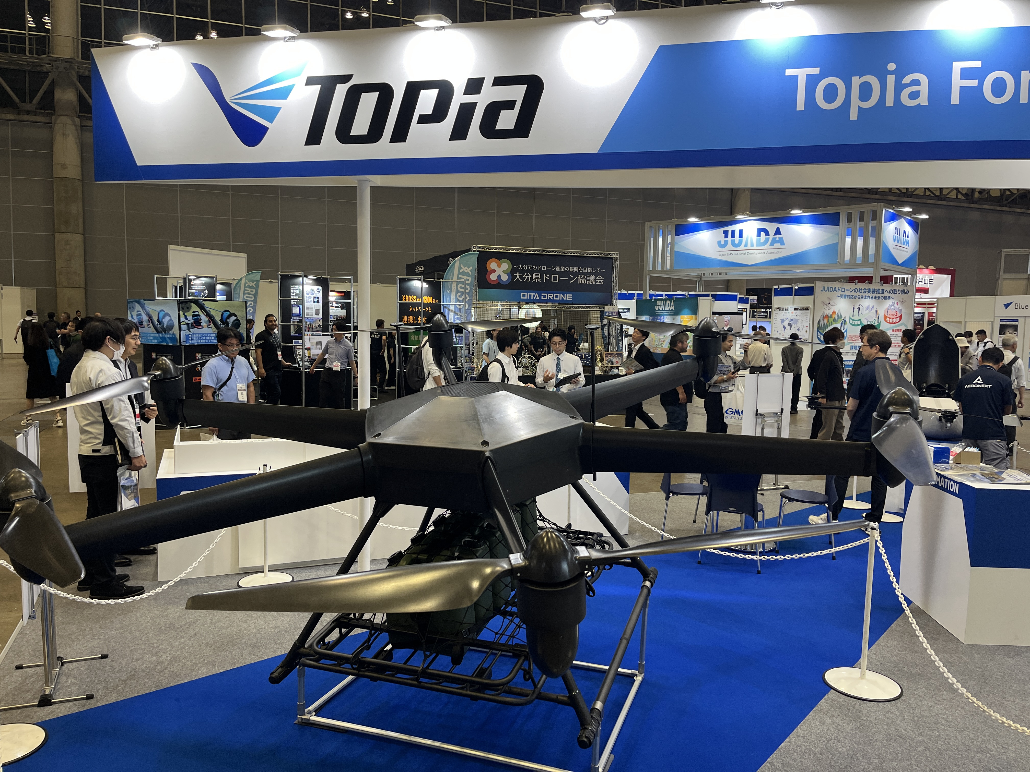 Exhibition hall with a large Topia banner overhead and people viewing drone displays below.