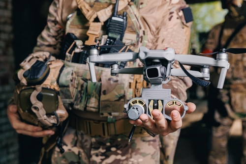 Person in camouflage holding a quadcopter drone with camera and a handheld remote controller, outdoors in tactical gear.