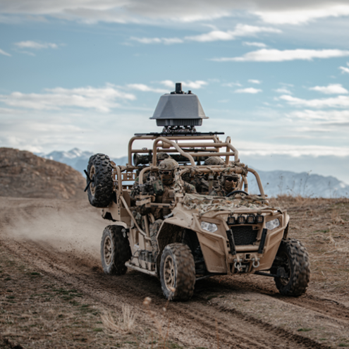 Dust-covered off-road vehicle with gear and two soldiers driving across a desert trail at speed.