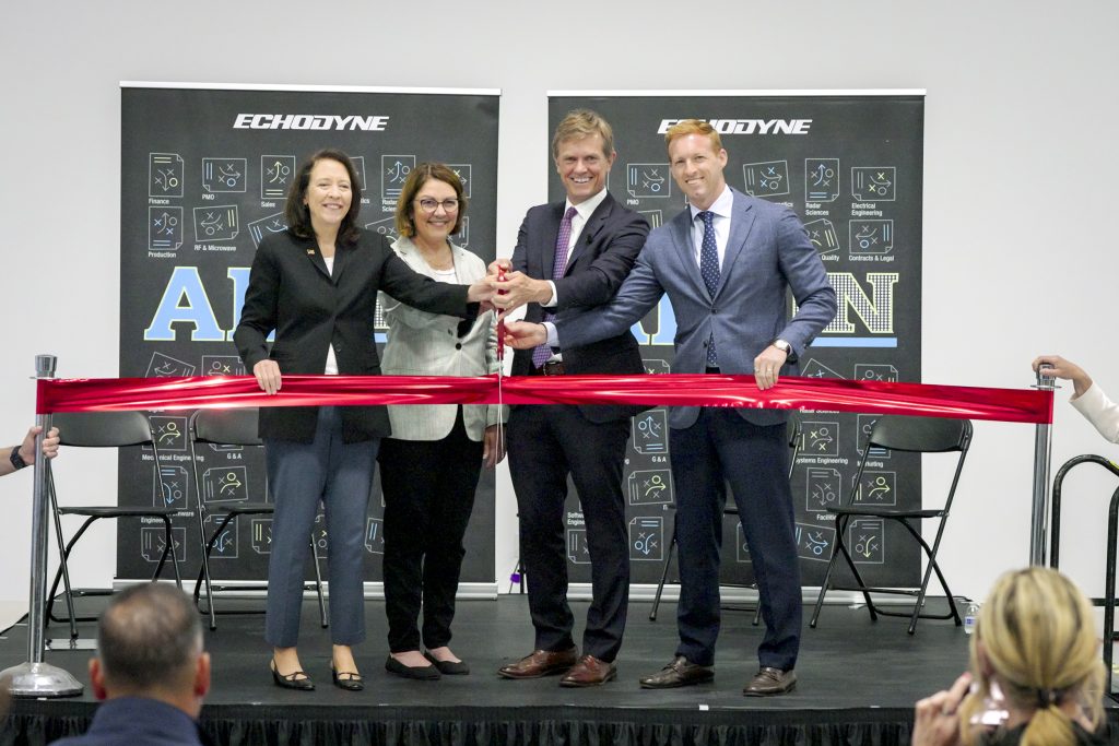 Four professionals in business attire cut a red ribbon at a corporate ribbon-cutting ceremony, with Echodyne banners in the background.