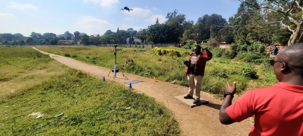 Man in red plaid shirt controls a drone from a dirt path, with a tripod-mounted device nearby as a drone flies overhead in a grassy field.
