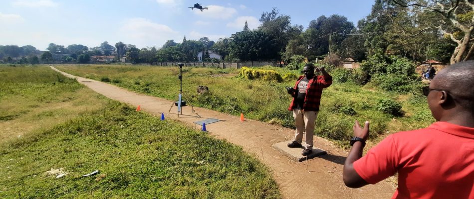 Man in red plaid shirt controls a drone from a dirt path, with a tripod-mounted device nearby as a drone flies overhead in a grassy field.