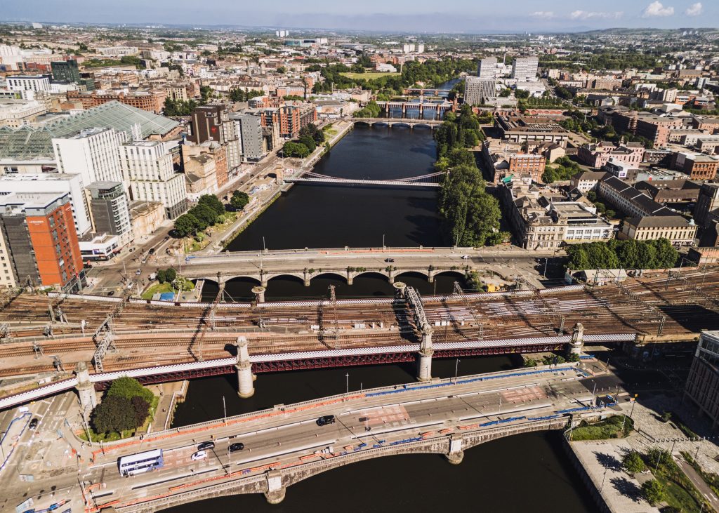 Aerial cityscape showing a wide river with several bridges, dense buildings along the banks, and railway tracks in the foreground.