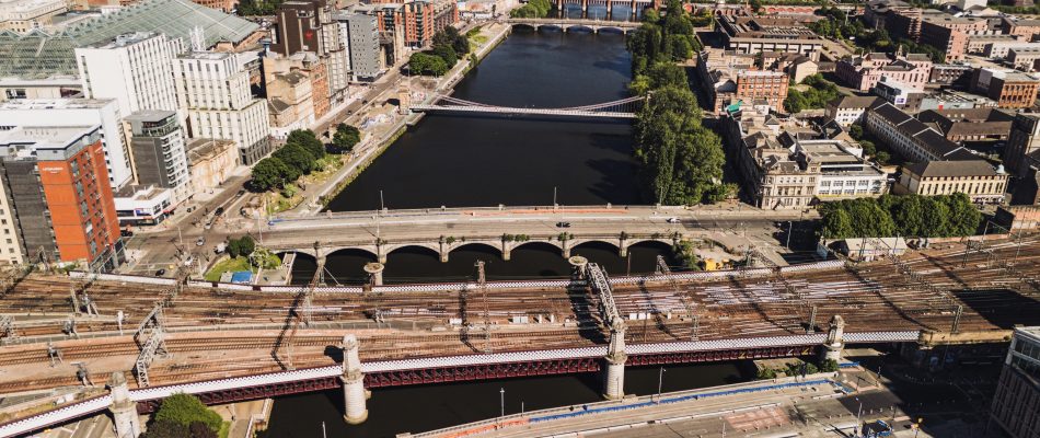 Aerial cityscape showing a wide river with several bridges, dense buildings along the banks, and railway tracks in the foreground.